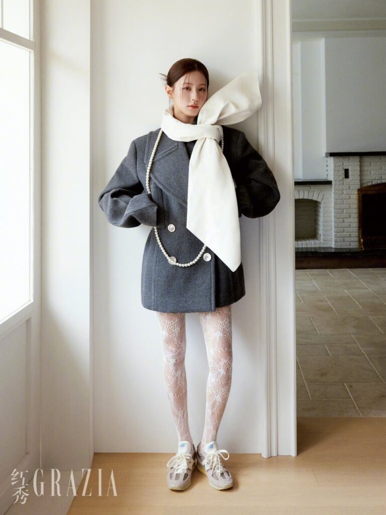 Miyeon leaning against white wall in gray coat and lace tights with dramatic ivory bow for Grazia