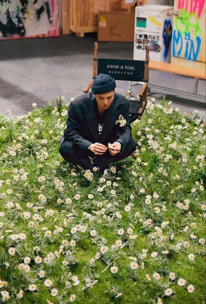 G-Dragon, dressed in a black suit and beanie, sits among blooming daisies while holding a flower, with a director's chair labeled 'Kwon Ji Yong' behind him