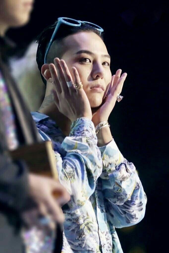 G-Dragon wearing a floral shirt and blue sunglasses on his head, striking a playful pose with both hands on his cheeks during a public appearance