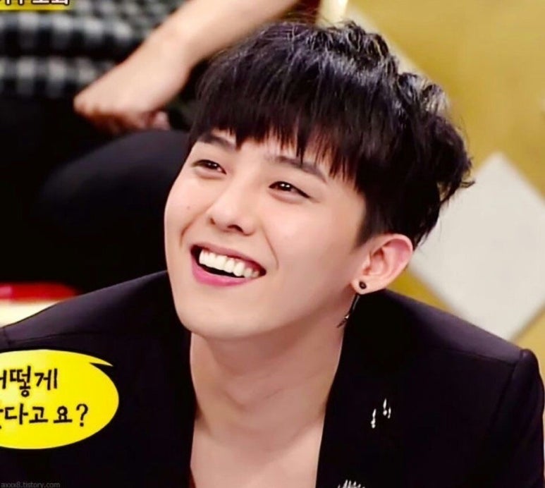 G-Dragon smiling brightly during a television show appearance, wearing a black jacket with a low neckline and small hoop earring