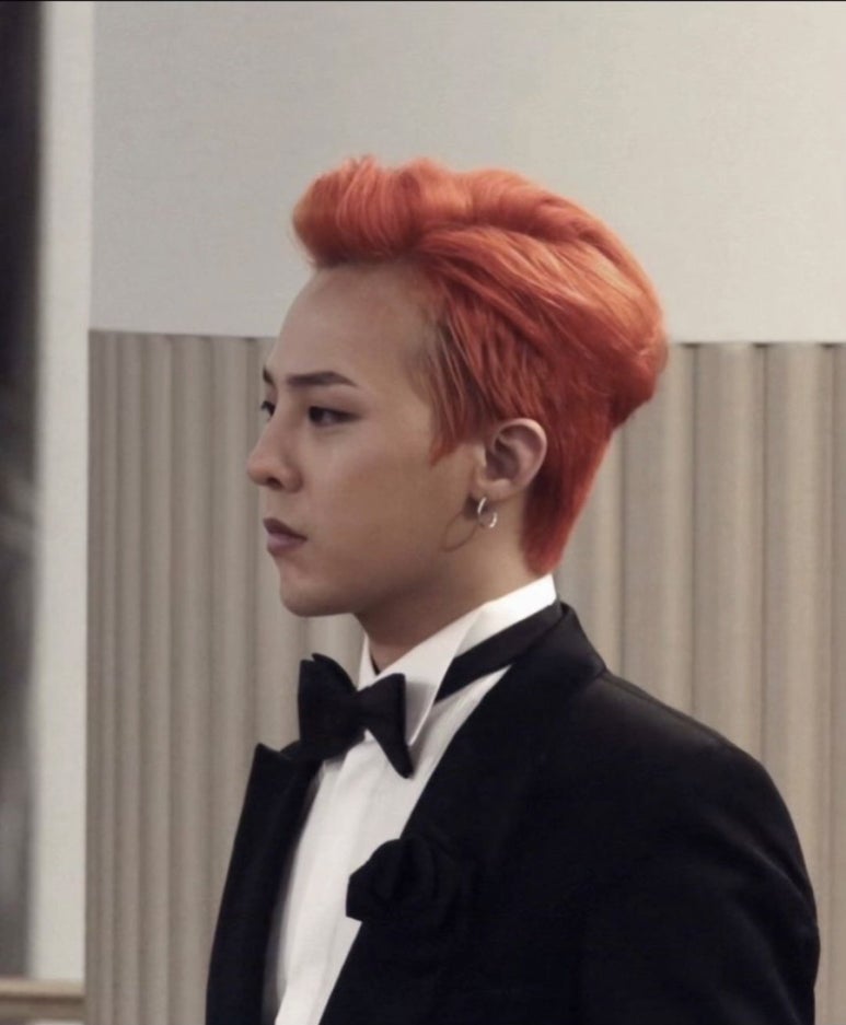 G-Dragon with vibrant orange hair, dressed in a black tuxedo with a bow tie and floral lapel pin, looking to the side in a formal setting