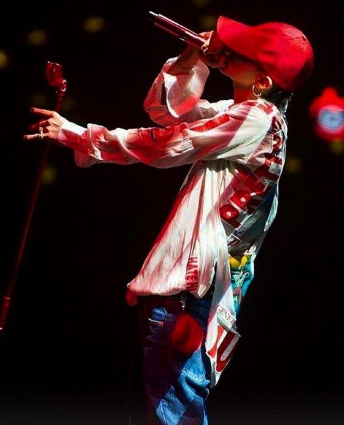 G-Dragon performing on stage in a red cap, patterned white shirt, and blue jeans, passionately singing into a microphone under dramatic red lighting