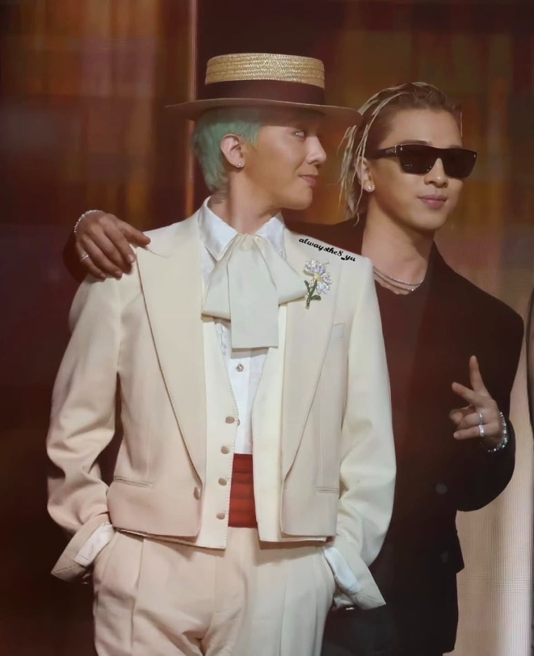 G-Dragon in a cream suit with a wide-brimmed straw hat and floral brooch standing beside Taeyang in a black outfit and sunglasses, posing together on stage