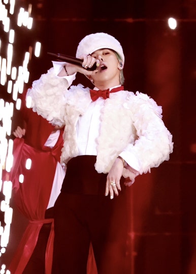 G-Dragon performing on stage in a white textured jacket, red bow tie, and matching white hat, singing into a microphone with dramatic stage lighting and sparks