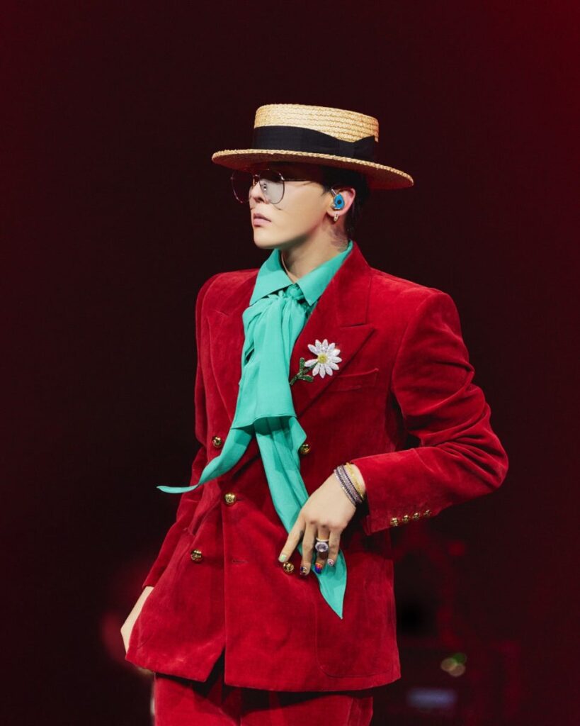G-Dragon wearing a bold red velvet suit with a turquoise silk scarf, straw boater hat, and round glasses, performing on stage with a daisy brooch pinned to his jacket.