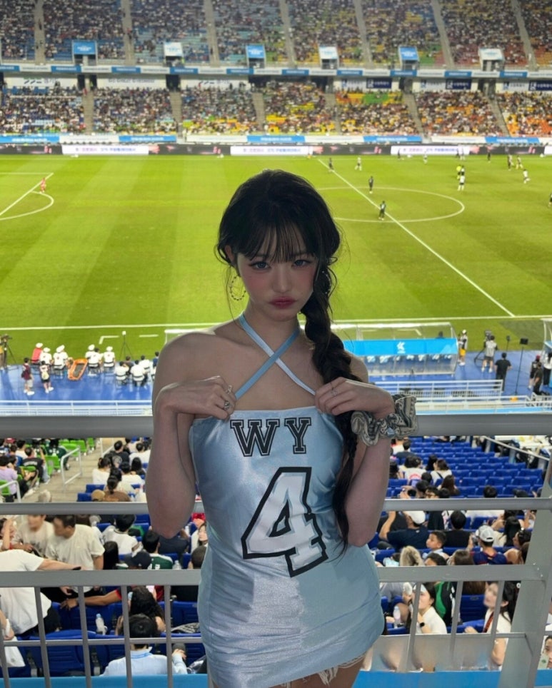 A young woman, Jang Wonyoung, is standing in a stadium wearing a light blue dress with a crisscross halter neck. The dress has the letters "WY" and the number "4" printed on it. She has her hair tied in a loose braid and is looking at the camera with a playful pout. A soccer field and a large crowd of spectators are visible in the background.