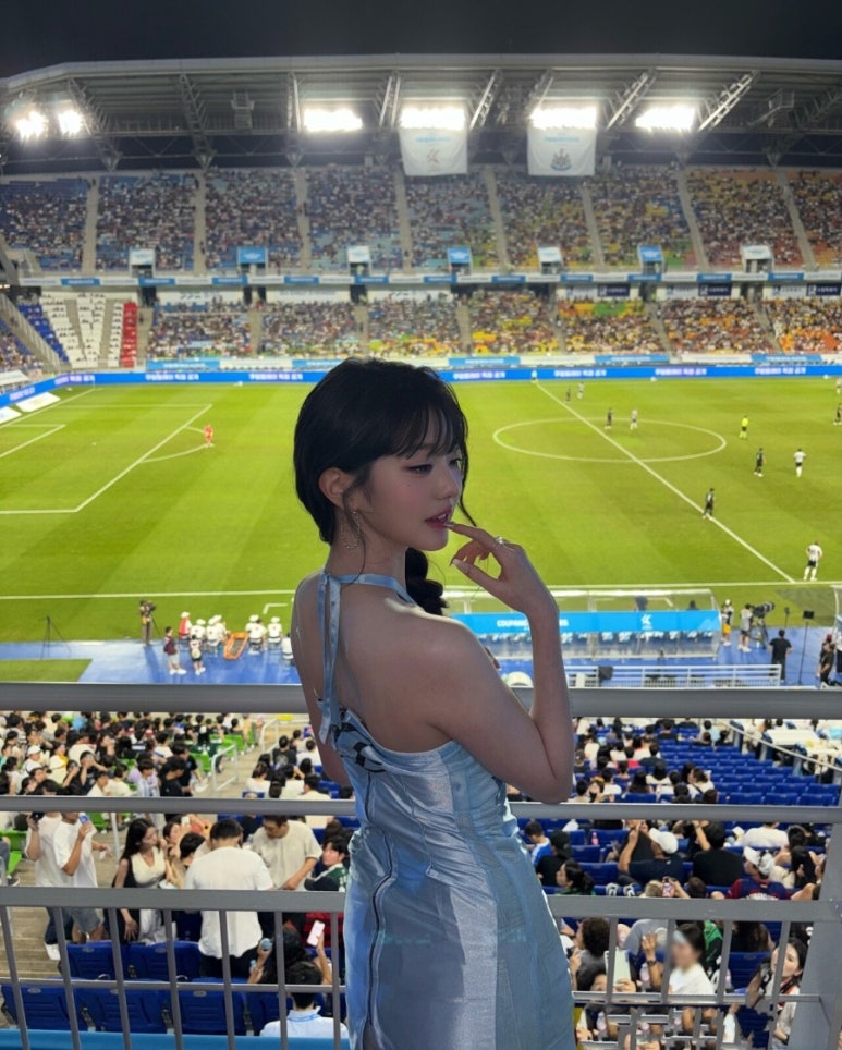 A young woman with dark hair and bangs, Jang Wonyoung, is seen from a slight side angle in a stadium. She is wearing a light blue, backless dress and is looking back over her shoulder towards the camera, with a finger touching her lips. The background shows a soccer field and a crowd of people in the stands.