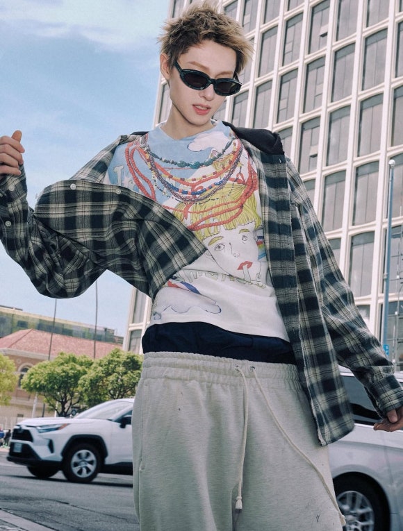 A young man with spiky blonde hair and sunglasses poses outdoors, wearing a graphic t-shirt and an open green plaid flannel shirt over a black hoodie.
