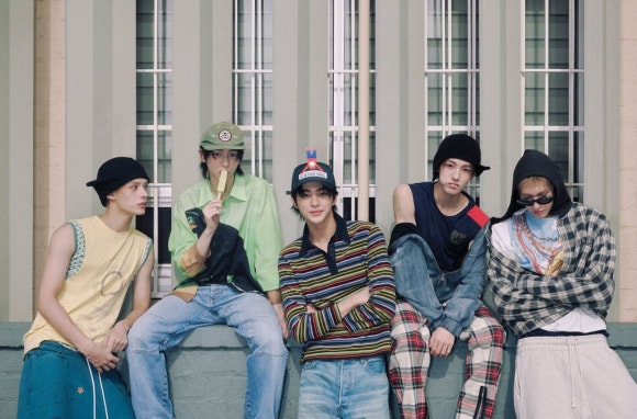 A group of five young men, CORTIS, is sitting in a row outdoors against a building with large windows. Each member wears a unique and stylish outfit with hats and accessories, showcasing a vibrant, street-style aesthetic.