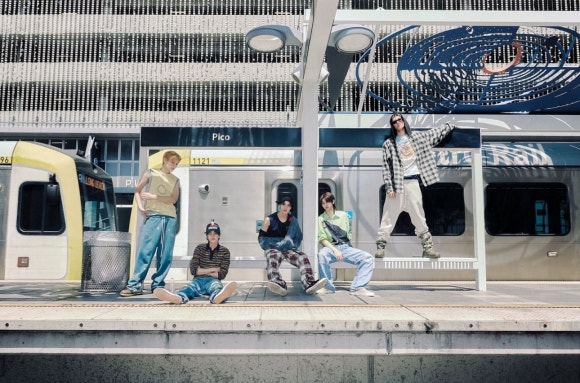 A five-member male group, CORTIS, is hanging out at a train station platform. They are posing in various positions—standing, sitting, and leaning—in front of a train, all wearing trendy, casual outfits. A sign for the "Pico" station is visible in the background.