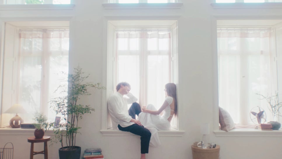 A man and woman dressed in white sit facing each other on a bright window sill, surrounded by soft natural light and minimalist decor