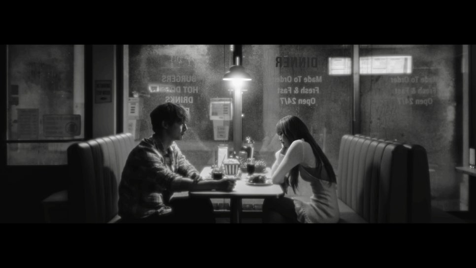 Black and white cinematic scene of a man and woman sitting across from each other in a diner booth on a rainy night, sharing a quiet, contemplative moment.