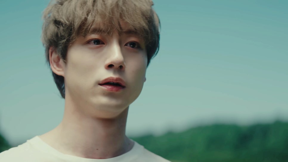 Close-up of a young man with light brown hair, wearing a white shirt, looking emotional under a clear blue sky with blurred greenery in the background