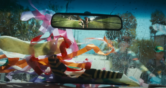 A point-of-view shot from inside a car's windshield, which is wet with rain or water droplets. Colorful streamers are taped to the dashboard, and the rearview mirror shows a reflection of a field where two people are visible.