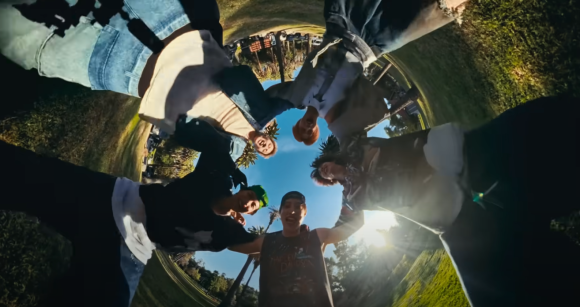 A low-angle, wide-angle shot of the five members of the group CORTIS looking down into the camera, forming a circle. The bright sun shines from behind them, and the background shows green grass and trees, creating a distorted, dynamic perspective.