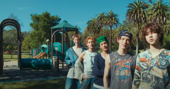 A five-member male group, CORTIS, standing together in a park with palm trees and a playground in the background under a clear blue sky. The members are wearing a variety of casual, street-style outfits.
