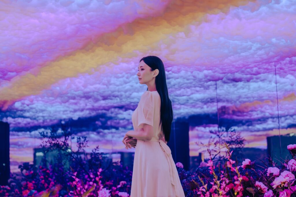 <em>A woman in a pastel dress stands in a dreamy digital flower garden under a colorful cloudscape at Waterworld Jeju, part of the visually stunning <strong>Waterworld Jeju discount</strong> attraction for families.</em>