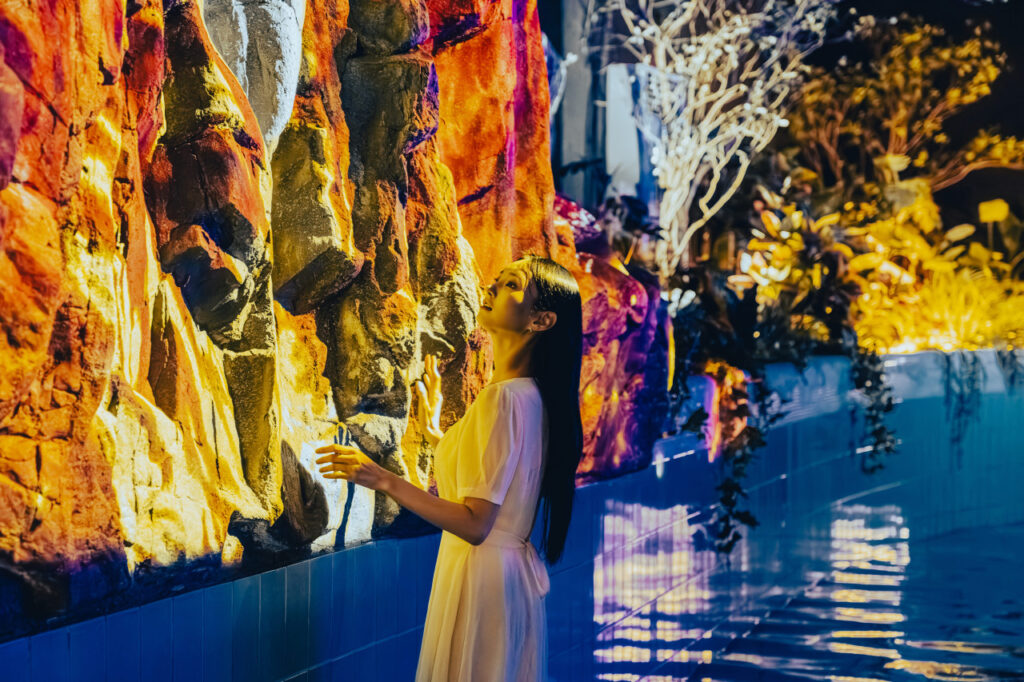 <em>A woman in a white dress explores a glowing volcanic rock installation surrounded by tropical plants at Waterworld Jeju, showcasing the immersive indoor experience available with <strong>Waterworld Jeju discount</strong> packages.</em>