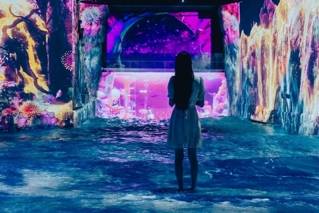 <em>A girl in a white dress stands barefoot in shallow water at Waterworld Jeju, surrounded by glowing digital art of waterfalls and underwater fantasy scenes — a magical indoor experience highlighting <strong>Waterworld Jeju discount</strong> benefits for families.</em>