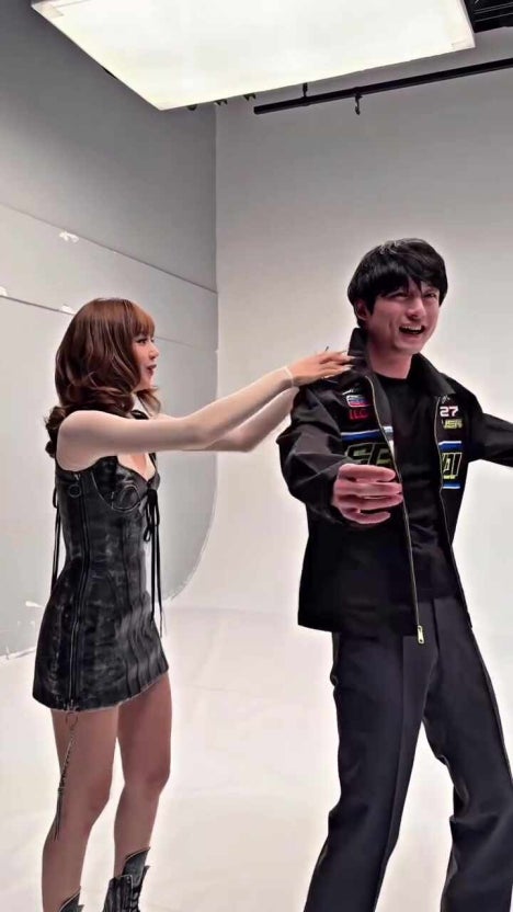 A woman in a black lace-up mini dress playfully interacts with a smiling man in a racing-style jacket during a studio photoshoot