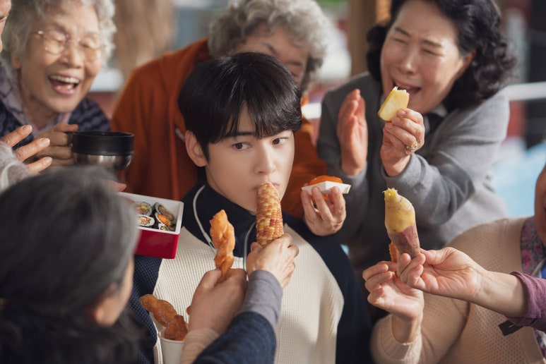 Cha Eun Woo surrounded by grandmothers offering food in a heartwarming scene from The First Ride
