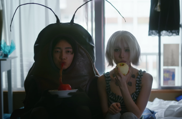 Sunmi eats an apple next to a person in a cockroach costume, creating surreal visual storytelling.
