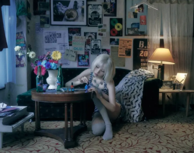 Sunmi paints her nails on a vintage carpet in a cluttered room, embracing an indie-rock aesthetic.