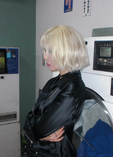 Side profile of Sunmi in a satin jacket, arms crossed, exuding edgy confidence in a laundromat.
