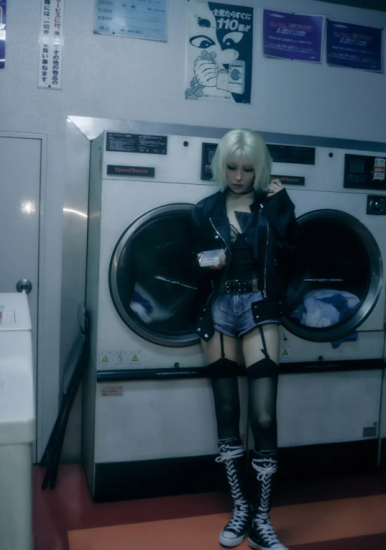 Sunmi stands in a laundromat wearing denim shorts and a leather jacket, embracing a bold punk look.