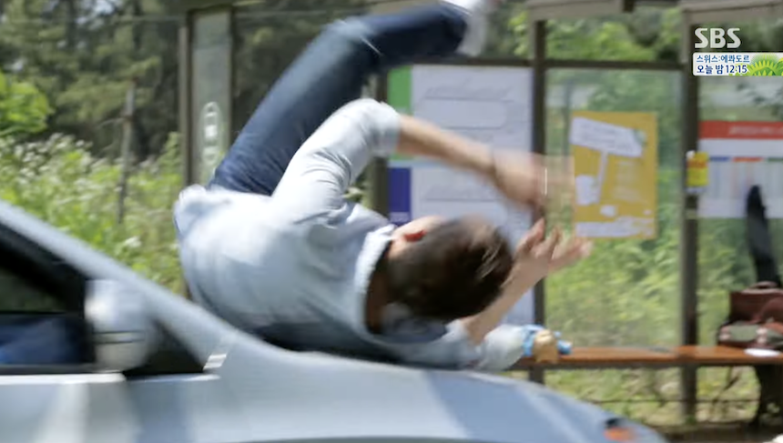A still image from a drama showing a stunt/action scene where a man is violently thrown over the hood of a light-colored car.