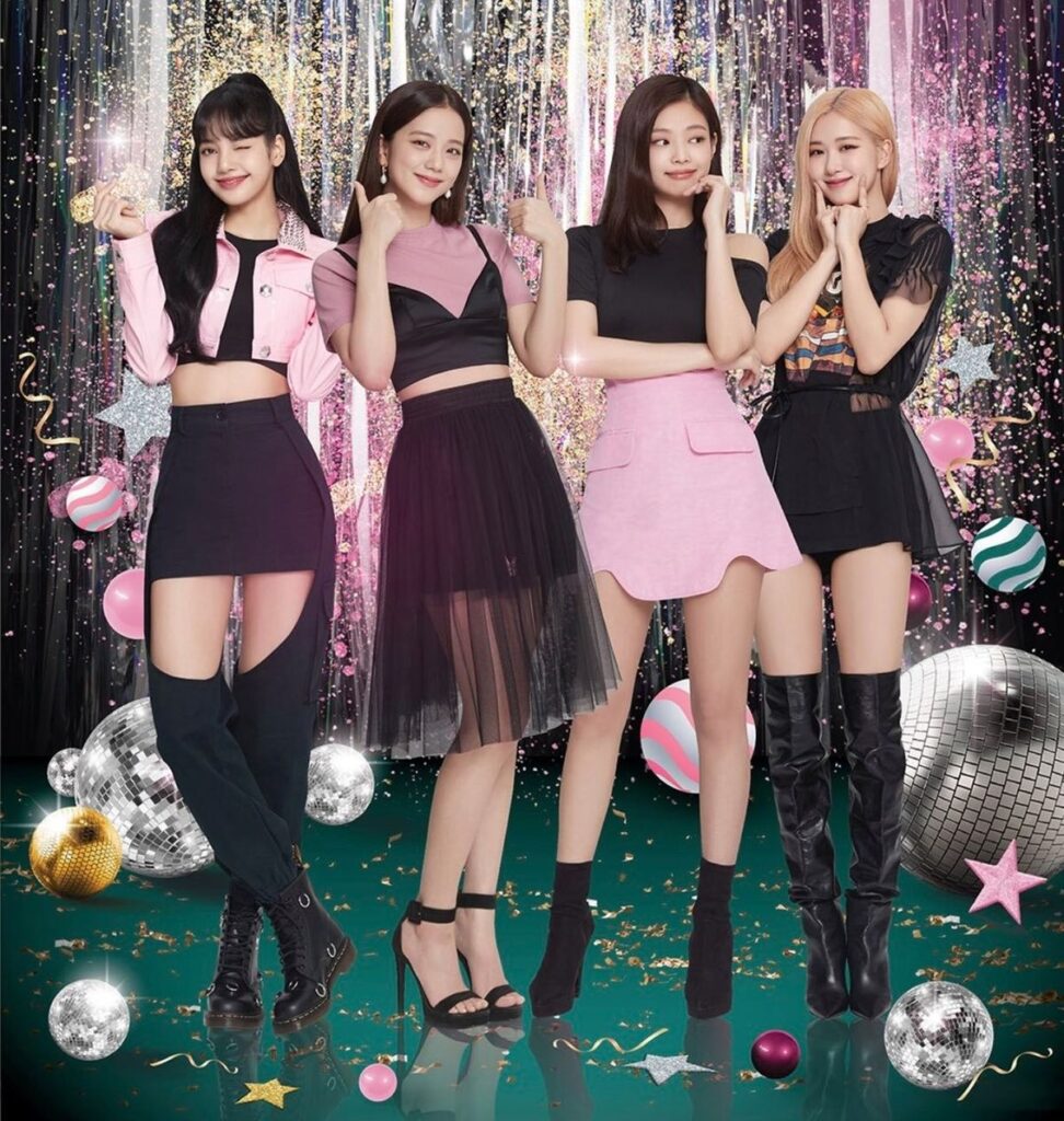 Vibrant party-themed photo of a girl group in black and pink stage outfits, surrounded by sparkling disco balls and colorful confetti decorations