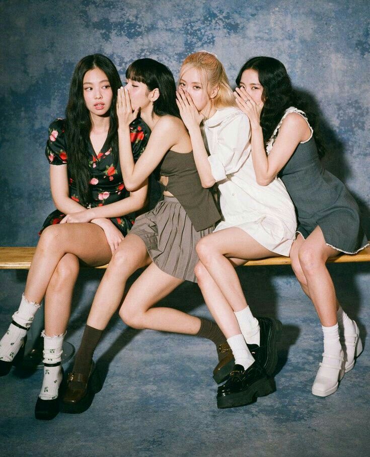 Four K-pop girl group members whispering secrets while sitting on a wooden bench, styled in casual retro fashion with a playful and candid vibe