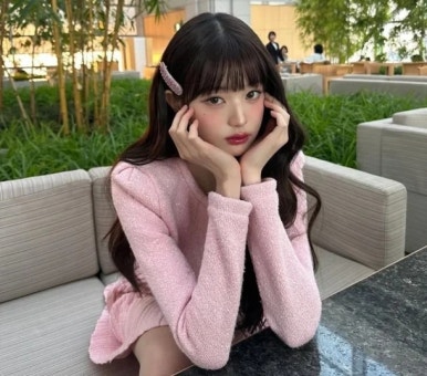 Jang Wonyoung wearing a soft pink long-sleeve dress with sparkly texture, sitting indoors with her hands on cheeks looking charming