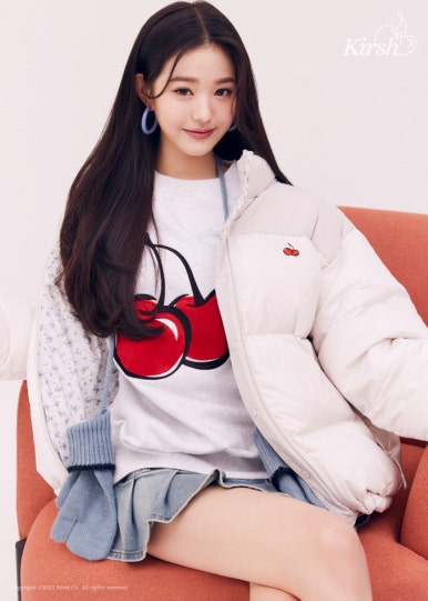 Jang Wonyoung modeling for Kirsh in a cherry-print tee, white padded jacket, and denim mini skirt sitting on an orange chair