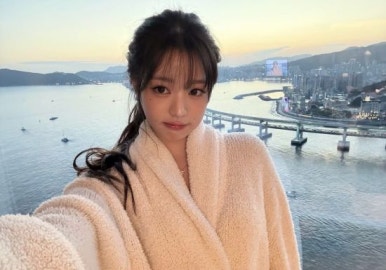 Jang Wonyoung taking a selfie in bathrobe in front of Gwangan Bridge view at sunset from a high-rise hotel room
