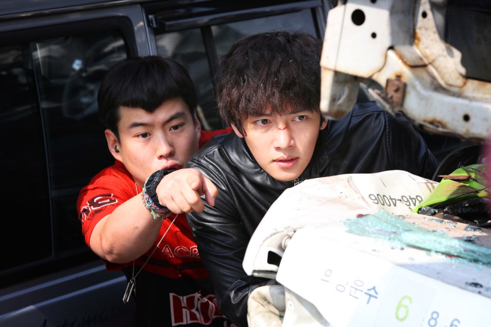 Two characters hiding behind a wrecked car in Fabricated City film